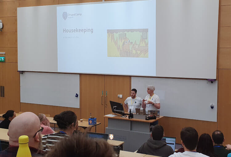 An audience of several people looking at Lewis and Rachel taking about housekeeping at the start of DrupalCamp England.