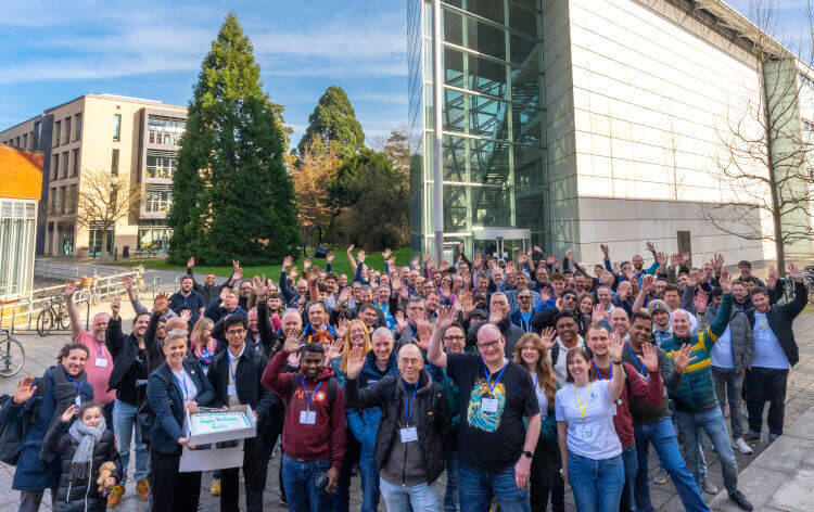 The group photo of DrupalCamp England 2025, with everyone waving. Stood outside the facilty of law campus at the university of Cambridge.